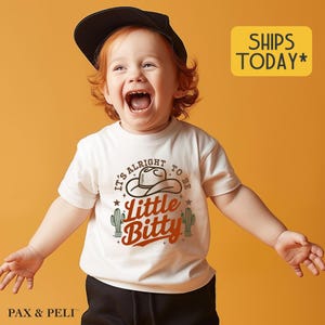 May include: A white t-shirt with a graphic design featuring a cowboy hat, cacti, and the text "It's alright to be little bitty". A young child is wearing the shirt and smiling.