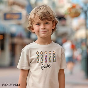 May include: A beige t-shirt featuring a colorful candle graphic and the word "five" in a handwritten font. The shirt is worn by a young child with blonde hair, smiling at the camera. The brand name "PAX & PELI" is printed at the bottom left.