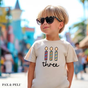 May include: A light beige t-shirt with a graphic of three colorful candles and the word "three" printed on the front.
