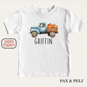 May include: White t-shirt featuring a light blue vintage truck carrying pumpkins. The name "GRIFFIN" is printed in black below the truck. The shirt is from PAX & PELI.