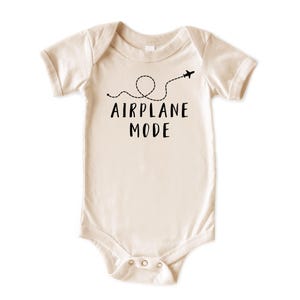 Puede incluir: Body de beb&eacute; color crema con mangas cortas. El texto negro "AIRPLANE MODE" est&aacute; impreso en la parte delantera, acompa&ntilde;ado de una l&iacute;nea de puntos y un peque&ntilde;o avi&oacute;n. El body tiene cierres de presi&oacute;n en la parte inferior.