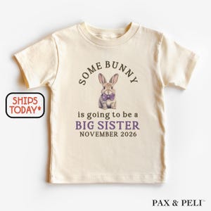 May include: Cream-colored toddler t-shirt with the text "SOME BUNNY is going to be a BIG SISTER NOVEMBER 2026". The shirt features a graphic of a bunny wearing a purple bow tie. The shirt is a short-sleeved crew neck.