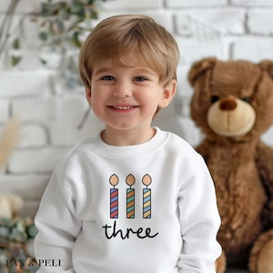 May include: A white sweatshirt with a colorful birthday candle graphic and the word "three" printed on the front.