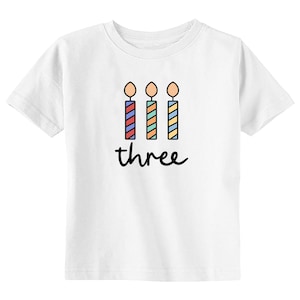 May include: White t-shirt with a graphic of three colorful candles and the word "three" printed on it.