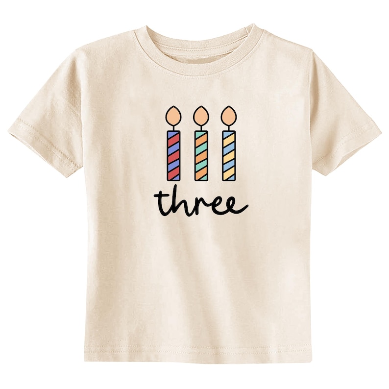 May include: A light beige t-shirt with a graphic of three colorful birthday candles and the word "three" printed on the front.