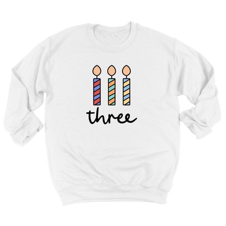 May include: White sweatshirt with a graphic of three colorful birthday candles and the word "three" printed below.