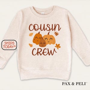 May include: A beige long-sleeved sweatshirt featuring the words "COUSIN CREW" in brown script. The design includes three smiling pumpkin illustrations and autumn leaves. The sweatshirt is a fall-themed garment.