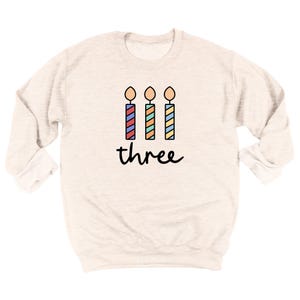 May include: A light beige sweatshirt with a graphic of three colorful birthday candles and the word "three" printed below.