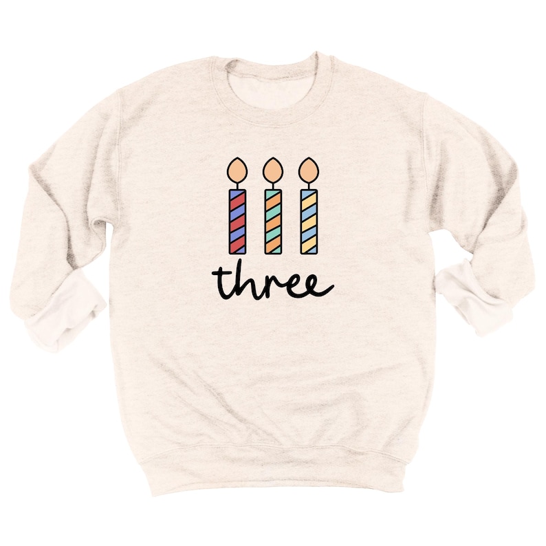 May include: A light beige sweatshirt with a graphic of three colorful birthday candles and the word "three" printed below.