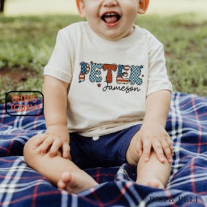 May include: A cream-colored t-shirt with the name "PETER Jameson" in red, white, and blue patriotic lettering. The child is wearing navy blue shorts and sitting on a blue and red plaid blanket.