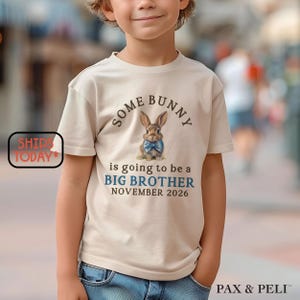 May include: A beige t-shirt with the text "SOME BUNNY is going to be a BIG BROTHER NOVEMBER 2026" and a watercolor illustration of a bunny wearing a blue bow tie. The shirt is worn by a child.