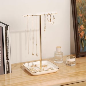 May include: A gold-toned jewelry stand with a square, white base and a vertical post. Several necklaces and earrings hang from the stand. A small perfume bottle and a jar with a gold lid sit nearby. The text "A compliment is something like a kiss through a veil. Hugo, Saint Denis" is visible.