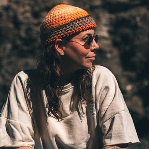 Könnte beinhalten: Eine Häkelmütze mit einem Farbverlauf in Orange-, Gelb- und Rosatönen sowie einem einfarbigen braunen Rand. Die Mütze wird mit einem cremefarbenen Kurzarm-T-Shirt kombiniert, das einen kleinen quadratischen Aufnäher auf der Brust mit teilweise sichtbarem Text hat.