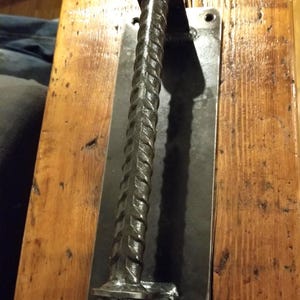 May include: A handcrafted, dark metal door pull with a textured, twisted bar design. The pull is mounted on a rectangular black metal plate, attached to a wooden surface. The hardware is designed for a rustic or industrial aesthetic.