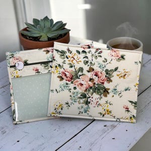 May include: Two floral fabric pouches with a cream background and a pattern of pink and yellow roses, green leaves, and blue accents. One pouch has a clear window and a zipper. A succulent plant in a terracotta pot is in the background.