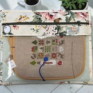 May include: A clear, zippered project bag with a floral print border, holding a cross-stitch project in a wooden hoop. The cross-stitch design features festive holiday motifs. A blue thread and a tag are visible.