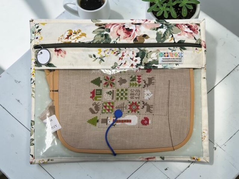 May include: A clear, zippered project bag with a floral print border, holding a cross-stitch project in a wooden hoop. The cross-stitch design features festive holiday motifs. A blue thread and a tag are visible.