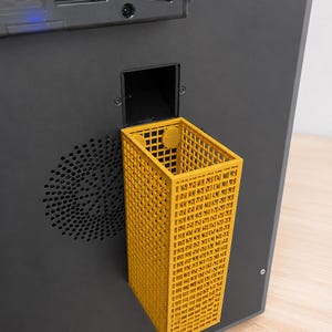 Könnte beinhalten: Ein rechteckiger gelber Kunststoffbehälter mit gitterartiger Struktur, der an der Seite eines dunkelgrauen elektronischen Geräts befestigt ist. Der Behälter verfügt über zwei kreisförmige Befestigungspunkte an der Oberkante und eine quadratische Öffnung.