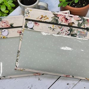 May include: Two clear, zippered pouches with a floral pattern border. The pouches have a light green interior with white polka dots. The floral pattern includes pink and cream roses and green leaves. Each pouch has a small, round tag attached to the zipper.