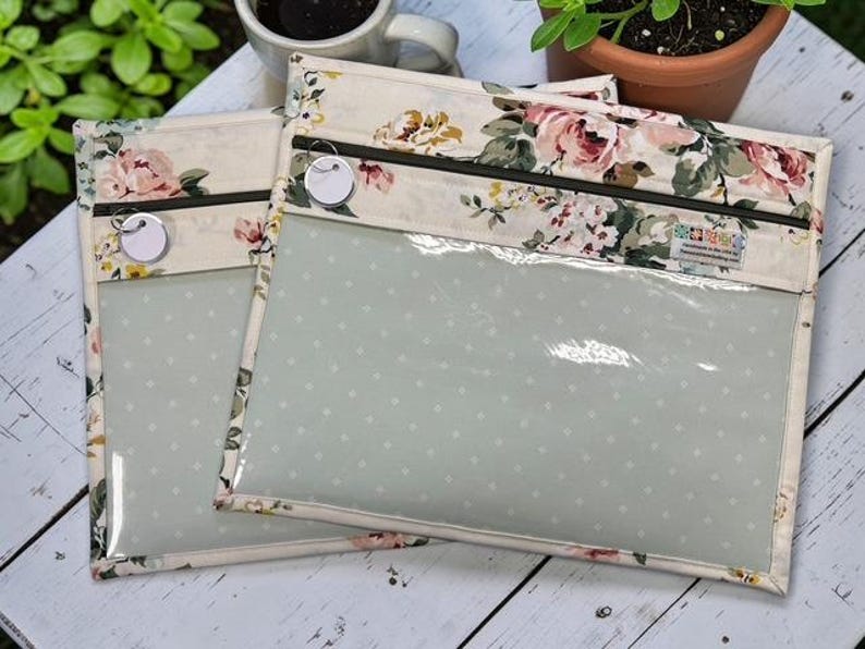 May include: Two clear, zippered pouches with a floral pattern border. The pouches have a light green interior with white polka dots. The floral pattern includes pink and cream roses and green leaves. Each pouch has a small, round tag attached to the zipper.