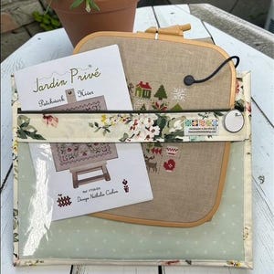 May include: A clear, floral-patterned pouch holds a cross-stitch project in a wooden hoop, with a partially visible design of a house and trees. A card with the text "Jardin Priv&eacute;" is also inside. A potted plant sits in the background.