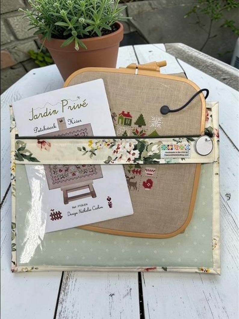 May include: A clear, floral-patterned pouch holds a cross-stitch project in a wooden hoop, with a partially visible design of a house and trees. A card with the text "Jardin Priv&eacute;" is also inside. A potted plant sits in the background.