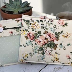 May include: Two fabric pouches with a floral pattern of pink, yellow, and blue roses on a cream background. One pouch has a clear pocket with a light blue polka dot pattern. A button and zipper closure are visible. A succulent plant in a terracotta pot and a cup of coffee are in the background.