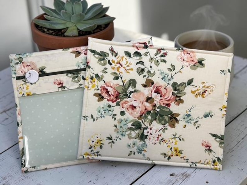 May include: Two fabric pouches with a floral pattern of pink, yellow, and blue roses on a cream background. One pouch has a clear pocket with a light blue polka dot pattern. A button and zipper closure are visible. A succulent plant in a terracotta pot and a cup of coffee are in the background.
