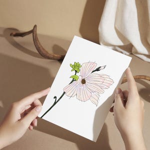 May include: A white card featuring a delicate illustration of a magnolia flower. The flower is depicted with soft pink petals and a green stem. The card is held by two hands, with a neutral background.