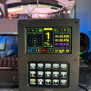 May include: A black carbon fiber racing dashboard with a digital display showing lap times, speed, and other performance metrics. It has multiple buttons for controlling various functions and a radar display. The screen displays the text "+0.457" and "12/12".
