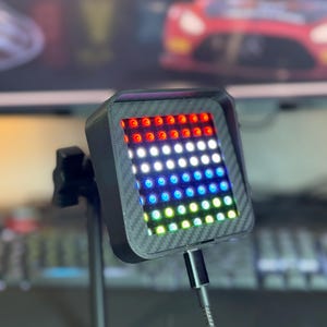 May include: A black and gray LED light panel with a carbon fiber texture. The panel is mounted on a flexible arm and has a USB cable.
