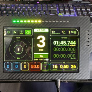 May include: A black digital display with a carbon fiber frame, showing various data points in green, yellow, and white. The screen displays lap times, fuel levels, and other performance metrics. The display is surrounded by a black keyboard.
