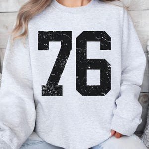 May include: A light gray sweatshirt with the number 76 printed in large, distressed black font across the front. The sweatshirt has long sleeves and a crew neck.