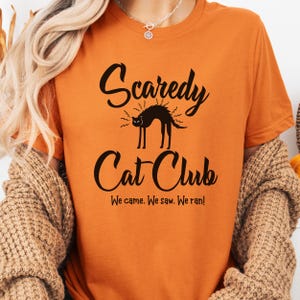 May include: Orange t-shirt with the text "Scaredy Cat Club" in black script, featuring a black cat illustration. The shirt also includes the phrase "We came. We saw. We ran!"