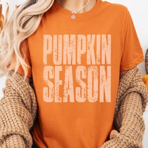 May include: An orange t-shirt with the words "PUMPKIN SEASON" printed in a light orange, distressed font. The shirt is a solid color and has short sleeves. The overall theme is fall and autumn.