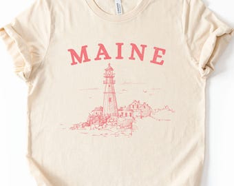 May include: A beige t-shirt with a pink graphic of a lighthouse and the word "MAINE" printed on the front.