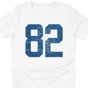 May include: White t-shirt with a large, distressed blue number 82 printed on the front. The number is centered and takes up a significant portion of the shirt's surface. The shirt has a classic crew neck design.