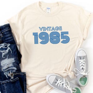 May include: A beige t-shirt with a blue vintage 1985 graphic. The shirt is laid flat on a white surface with a pair of gray sneakers.