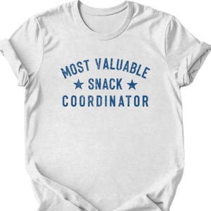 May include: White t-shirt with the blue text "MOST VALUABLE SNACK COORDINATOR" and two blue stars. The shirt has a crew neck and short sleeves. The text is in a distressed font.