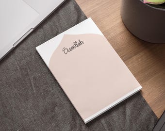 May include: A white notepad with a pink background and the word "Bismillah" written in black cursive. The notepad is on a brown wooden surface.