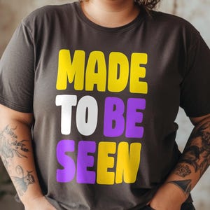 Könnte beinhalten: Dunkelgraues T-Shirt mit dem Text "MADE TO BE SEEN" in Gelb, Weiß und Lila. Das T-Shirt hat einen Rundhalsausschnitt.