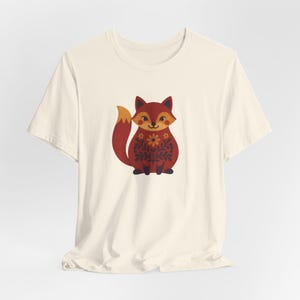 May include: A cream-coloured short-sleeve t-shirt featuring a centred graphic of a stylised fox. The fox is coloured in shades of red and orange with a dark brown floral pattern on its body, a bushy tail, and a small flower centred on its chest.
