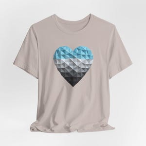 Puede incluir: Una camiseta de color beige claro con un diseño de corazón geométrico. El corazón está compuesto por formas triangulares en tonos de azul, gris y negro. La camiseta tiene mangas cortas y cuello redondo.