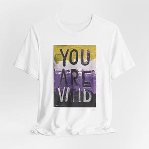 May include: White t-shirt with a graphic print of the words "YOU ARE VALID" in purple, yellow, and black paint. The graphic is in a rectangular frame.