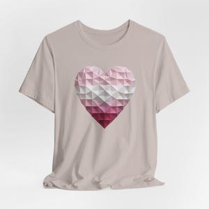 May include: A light pink t-shirt featuring a geometric heart design. The heart is composed of triangular shapes in shades of pink, white, and burgundy. The shirt has short sleeves and a crew neck.