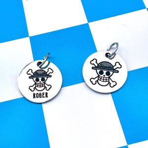 Può includere: Due targhette circolari in metallo color argento con un'incisione nera raffigurante un teschio con ossa incrociate che indossa un cappello di paglia. Una targhetta riporta il nome ROGER in lettere maiuscole nere. Ogni targhetta ha un anellino.