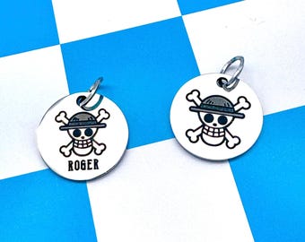 May include: Two circular silver-toned metal tags featuring a black engraved skull and crossbones design wearing a straw hat. One tag includes the name ROGER printed in black capital letters below the graphic. Each tag has a small metal jump ring.