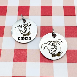 May include: Two circular silver-toned metal tags featuring a black engraved illustration of the character Gonzo with a bow tie. One tag includes the text GONZO in a bold, blocky font. Each tag has a small metal jump ring attached at the top.