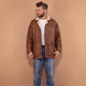 May include: A brown leather jacket with a cream-colored shearling collar and lining. The jacket has a button-up front, two front pockets, and a classic, vintage style. The person is wearing blue jeans and brown shoes.