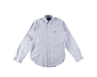 May include: Light blue button-down shirt with long sleeves. The shirt features a button-down collar and a small, dark blue embroidered logo on the chest. The shirt is made of a lightweight fabric and has a curved hemline.